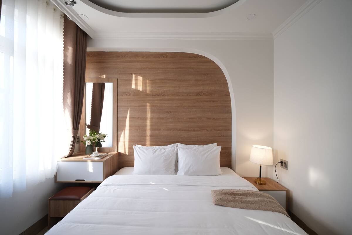 Habitación de hotel con una cama grande, cabecera de madera, mesita de noche con lámpara y un pequeño banco.