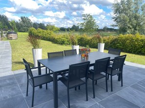 Une terrasse avec une table rectangulaire et six chaises, entourée de plantes en pot et offrant une vue sur un champ verdoyant et des arbres.