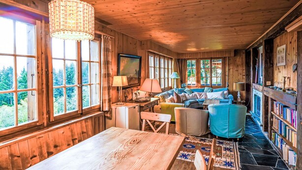 A cozy cabin interior with wooden walls, a large window overlooking a forest, a blue sofa, and a bookshelf filled with books. - Beautiful private villa for 6 guests with WIFI, TV, terrace and panoramic view (Les Collons)