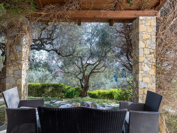 A covered outdoor seating area with stone pillars, wicker furniture, and a view of trees and greenery.