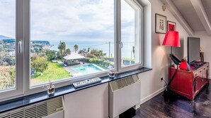 Habitación con un ventanal grande que da a una piscina y una vista panorámica del mar y las colinas.