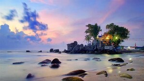 Una playa con rocas y un edificio con luces, palmeras y una puesta de sol.