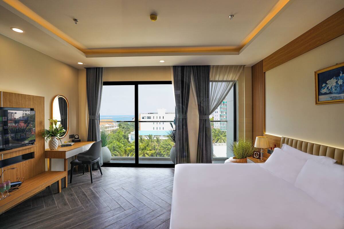 Une chambre d’hôtel avec un grand lit, un bureau avec une chaise, une télévision et une vue sur la ville grâce à de grandes fenêtres.
