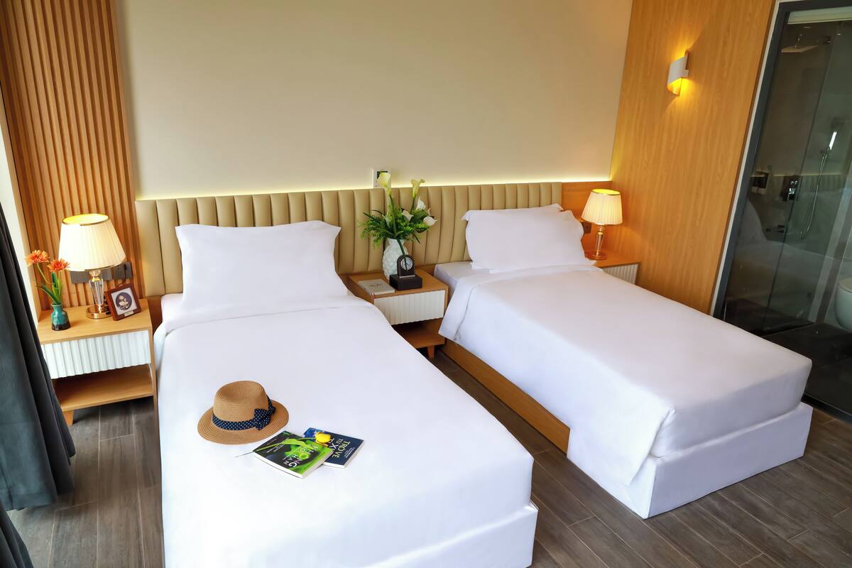 Une chambre d’hôtel avec deux lits simples, des tables de chevet avec des lampes, un vase avec des fleurs et une tête de lit en bois.