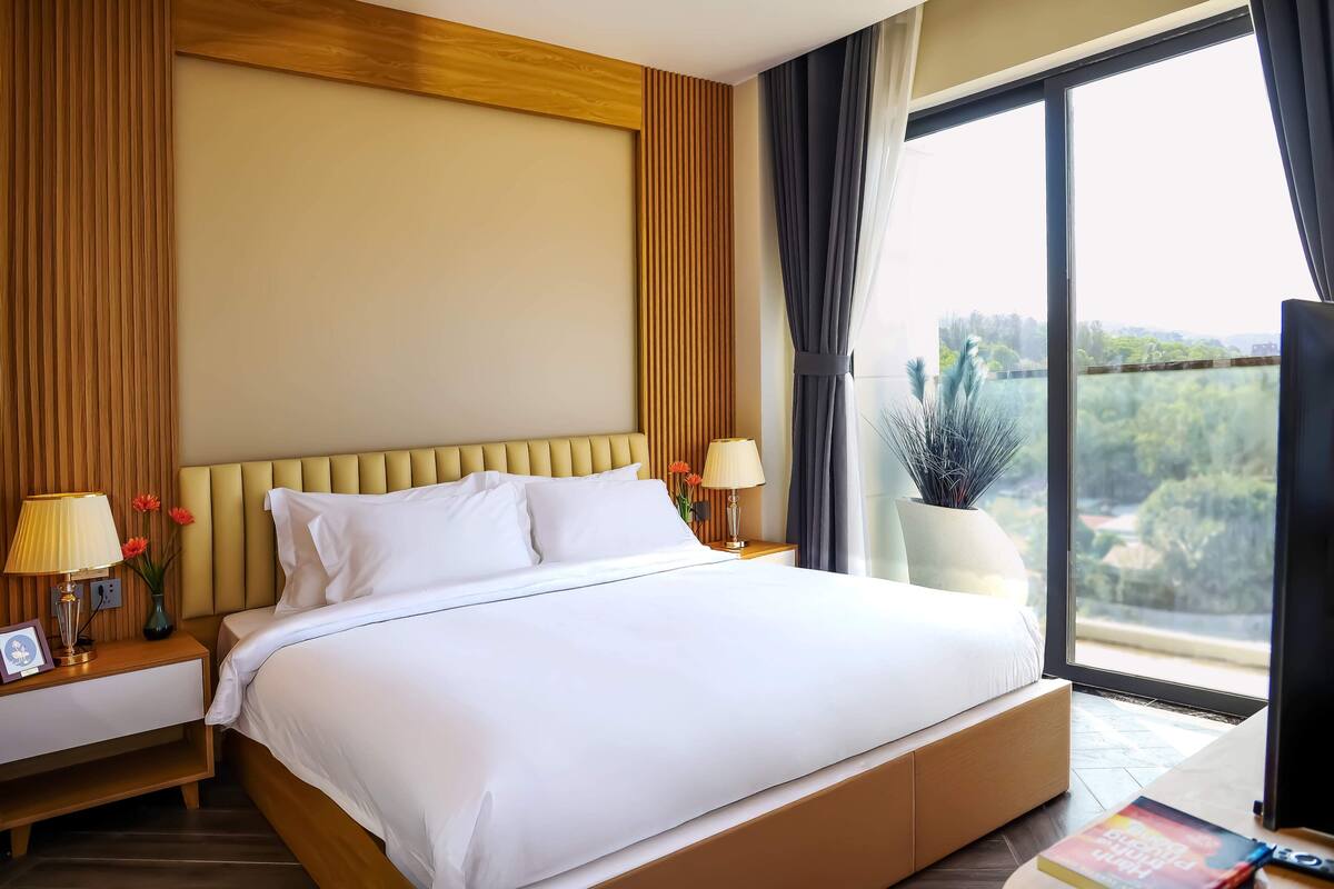 Une chambre d’hôtel avec un grand lit, une table de chevet avec une lampe, une plante en pot et une vue sur la verdure par la fenêtre.