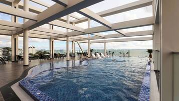 An indoor swimming pool with clear blue water, surrounded by white pillars and a view of the sea.