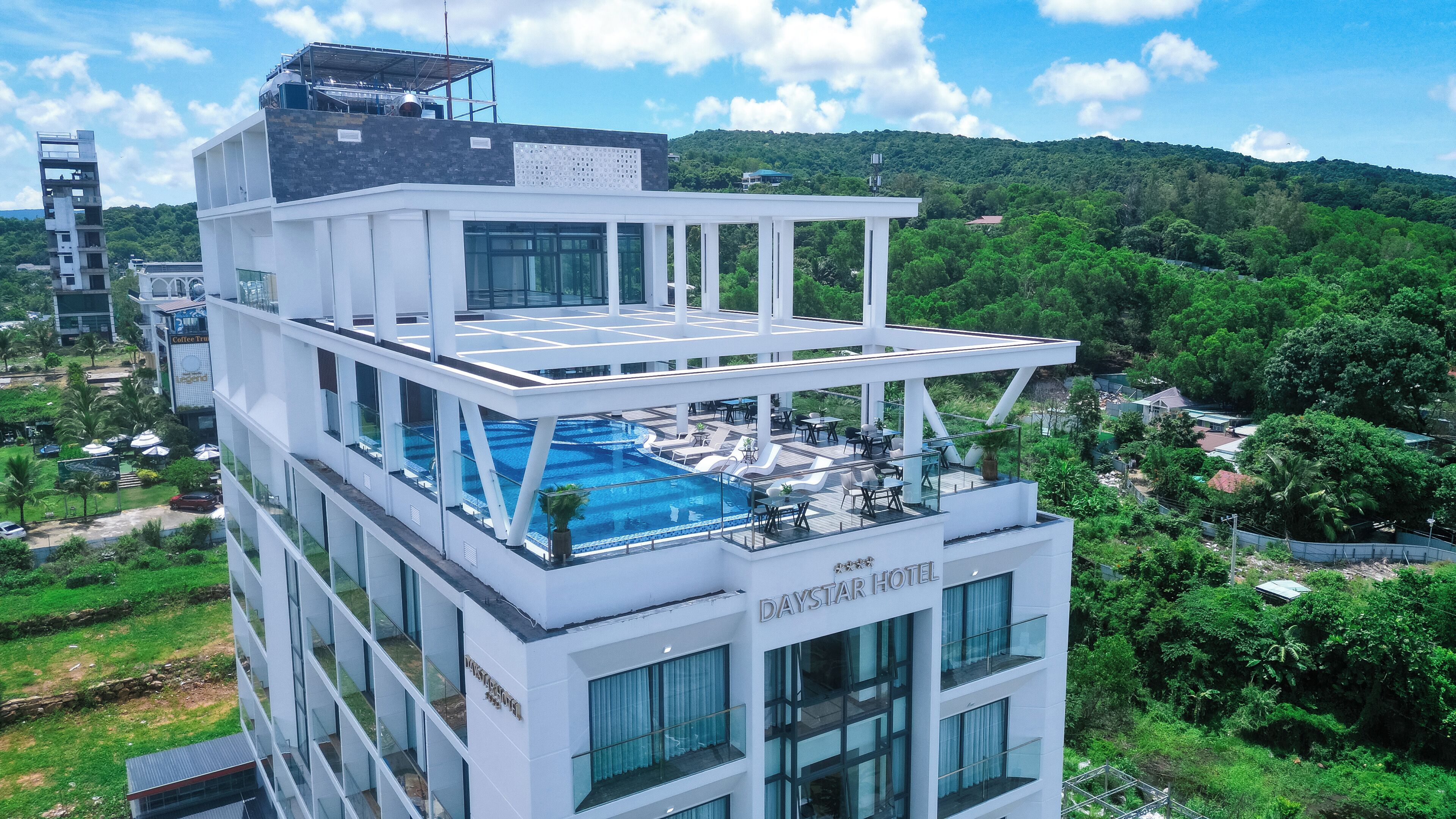 A multi-story hotel with a rooftop pool, large windows, and a view of the surrounding greenery.