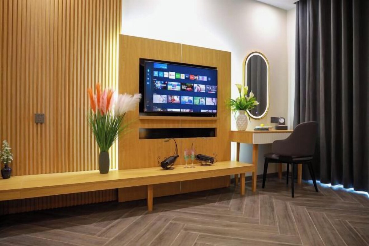 A modern hotel lobby with a wooden reception desk, a flat-screen TV mounted on the wall, a mirror, and a seating area with a bench.