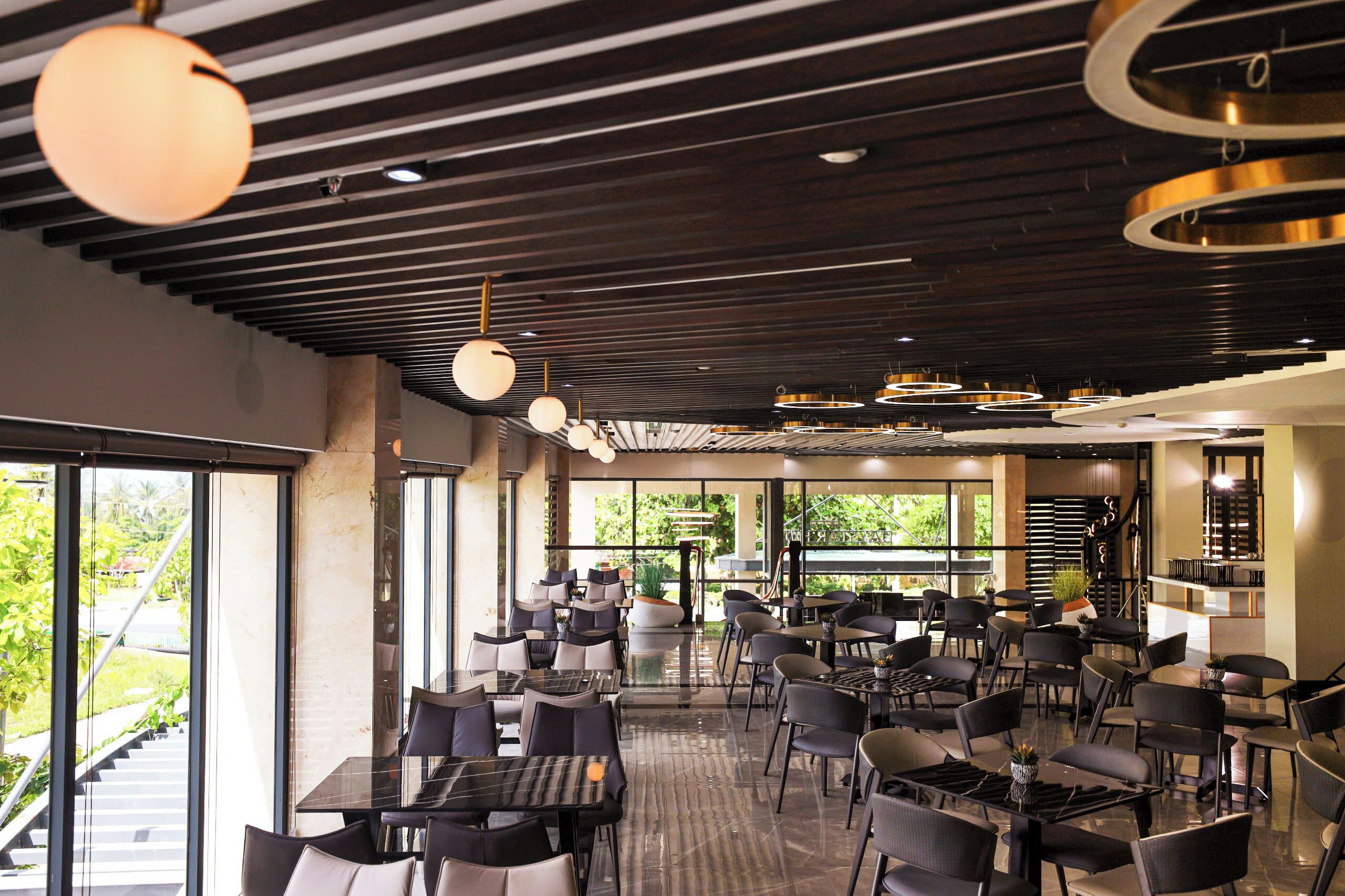 A spacious indoor area with multiple tables and chairs, large windows offering views of greenery, and modern lighting fixtures.