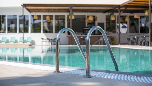 A swimming pool with metal railings, a covered seating area with chairs, and a building with large windows in the background. - Lime Hotel Faliraki (Faliraki)