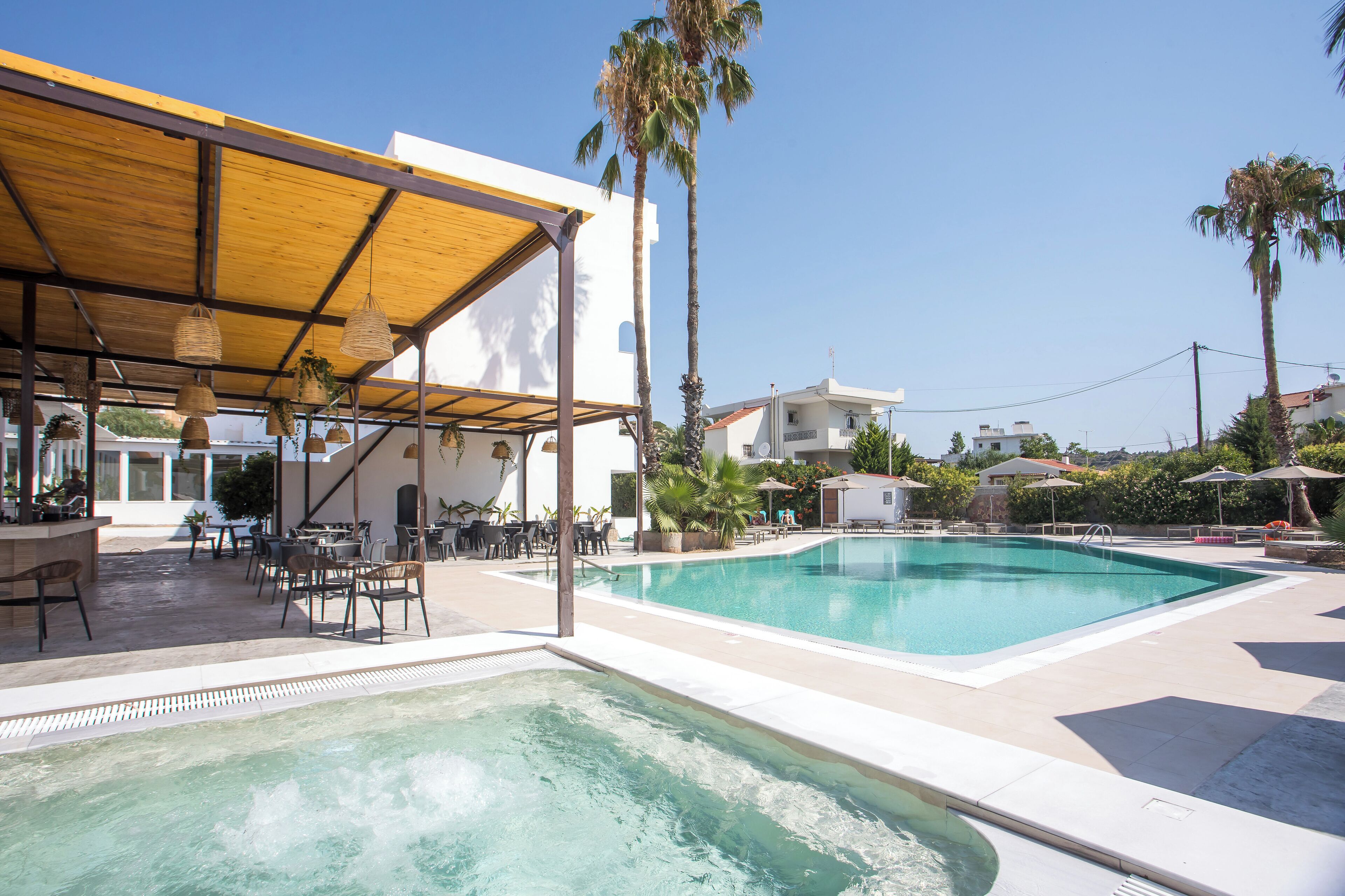 Zona de piscina con área de estar techada, palmeras y cielo azul despejado.