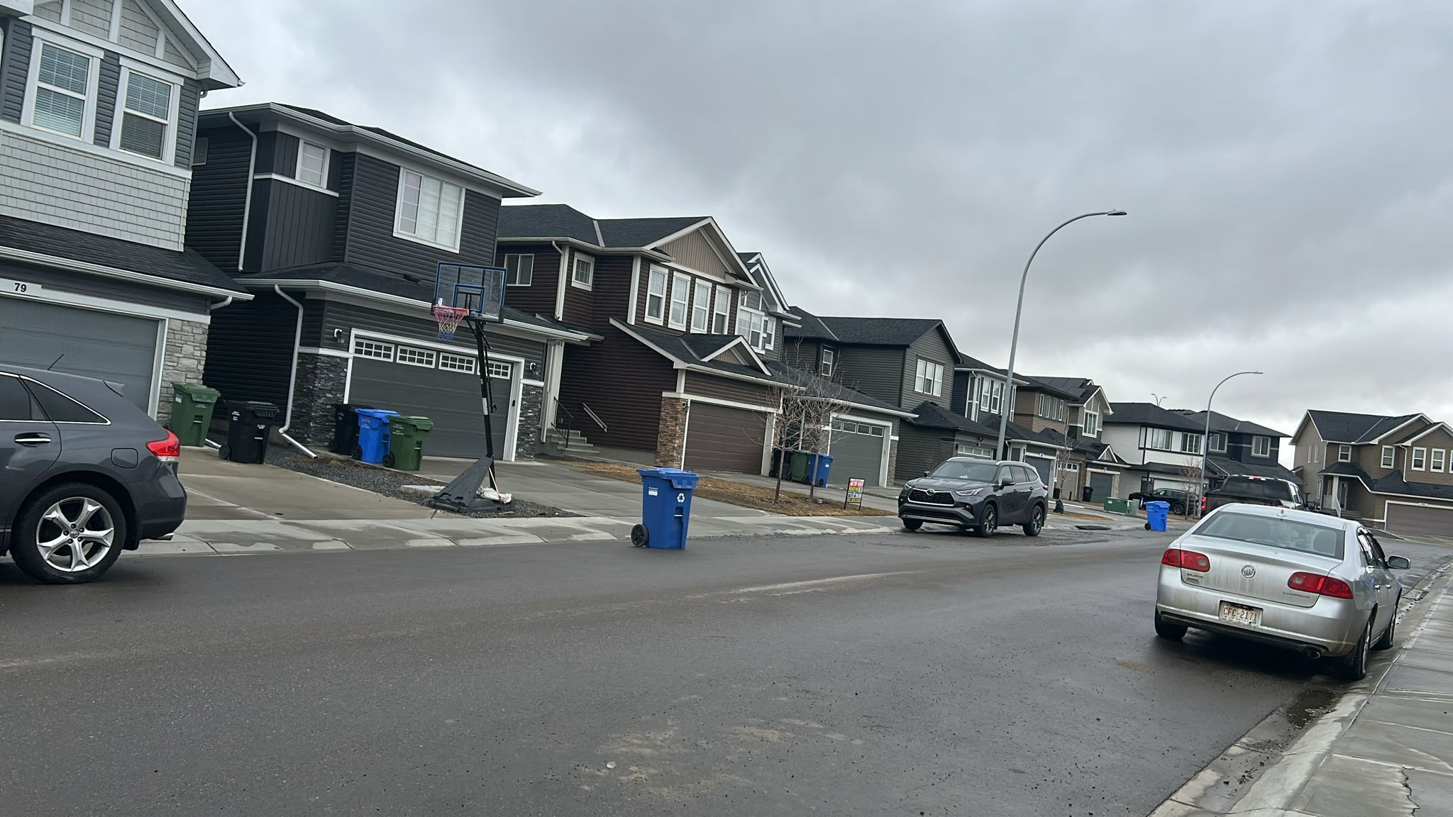A residential street with houses, parked cars, and a basketball hoop in the driveway.