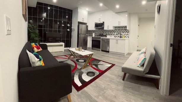 Ein modernes Wohnzimmer mit einer schwarzen Couch, einem weißen Sessel und einem Couchtisch. Das Zimmer verfügt über eine Küche mit weißen Schränken und einer schwarzen Spritzschutzwand.