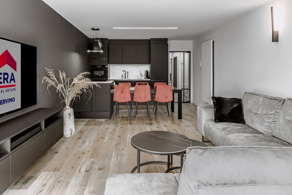 A modern living room with a grey sofa, a small round table, and a TV on a wall-mounted shelf. There is a kitchen area in the background with dark cabinets and a dining table with four chairs.