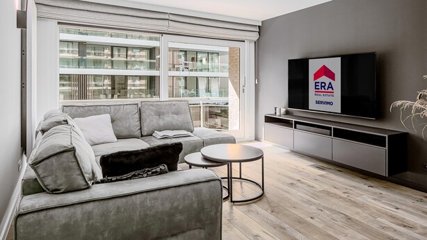 Ein modernes Wohnzimmer mit einem grauen Sofa, einem kleinen runden Tisch und einem an der Wand befestigten Flachbildfernseher.