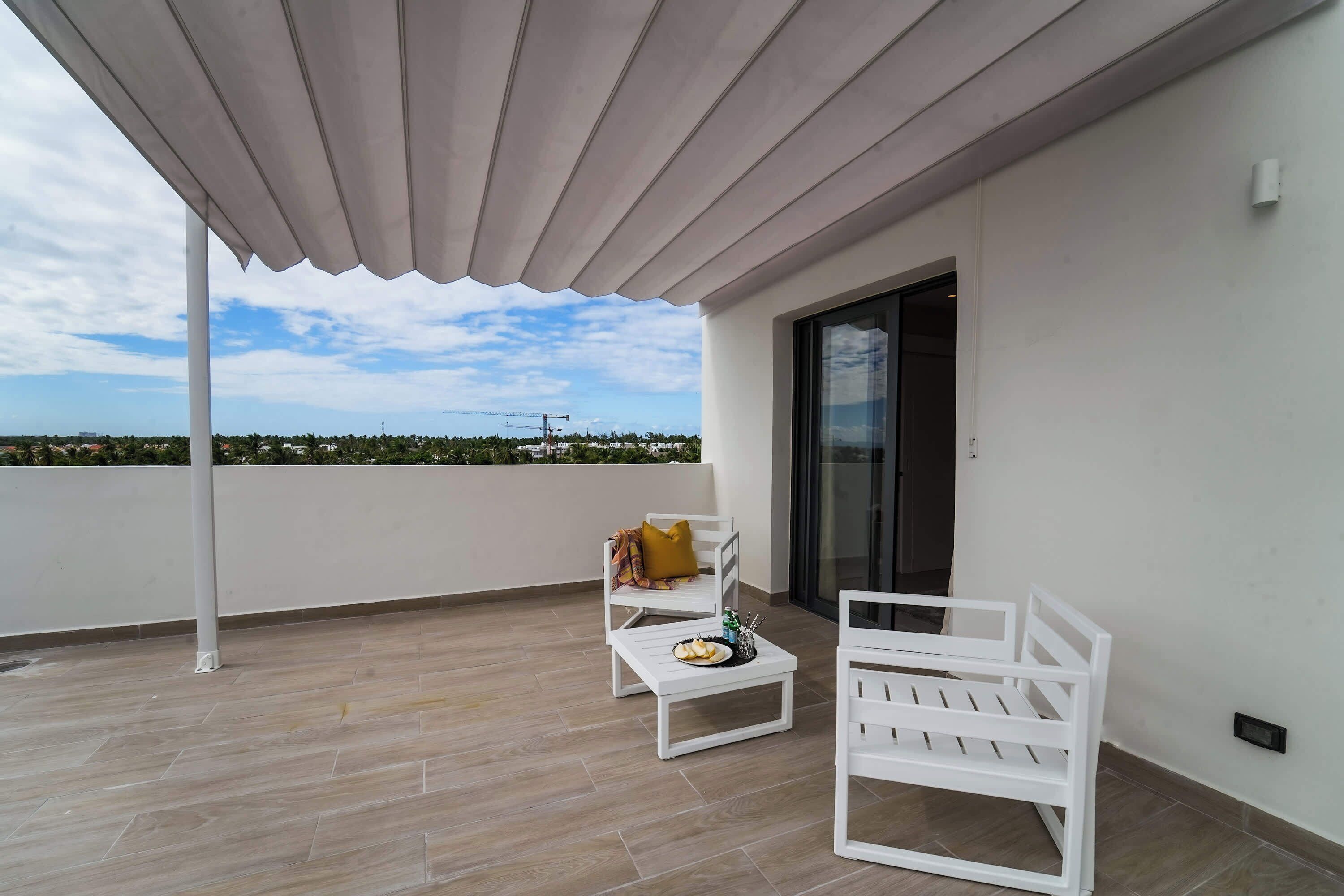 Een balkon met een wit plafond, twee witte stoelen, een klein tafeltje met een bord eten en een geel kussen.