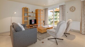 A modern living room with a grey sofa, a white armchair, a wooden cabinet, a television, and a view of the outside balcony. - Apollo II 0203 met garage (NIEUWPOORT)