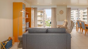 A modern living room with a grey sofa, a wooden cabinet, a floor lamp, and a view of the city through large windows. - Apollo II 0203 met garage (NIEUWPOORT)