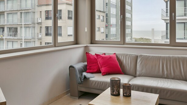 Ein modernes Apartment mit einem beigefarbenen Sofa, roten Kissen und zwei dekorativen Vasen auf einem Holztisch. Große Fenster bieten Blick auf eine Stadtlandschaft.