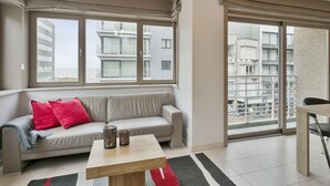 Ein modernes Wohnzimmer mit einer grauen Couch, roten Kissen, einem Holztisch und großen Fenstern mit Blick auf eine Stadtlandschaft.