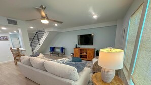 A modern living room with a sofa, a TV, a ceiling fan, and a staircase.