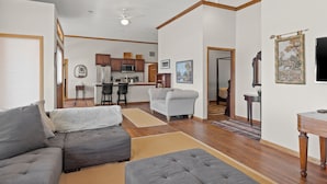 A spacious living area with a gray sectional sofa, a white armchair, and a wooden coffee table. A kitchen with dark wooden cabinets and a white refrigerator is visible in the background.