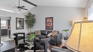 A modern living room with a black leather sofa, a glass coffee table, a black armchair, a small round table, and a floor lamp. There is a ceiling fan, a potted plant, and a poster on the wall. - Unwind in this newly updated 4-bedroom home (Laramie)