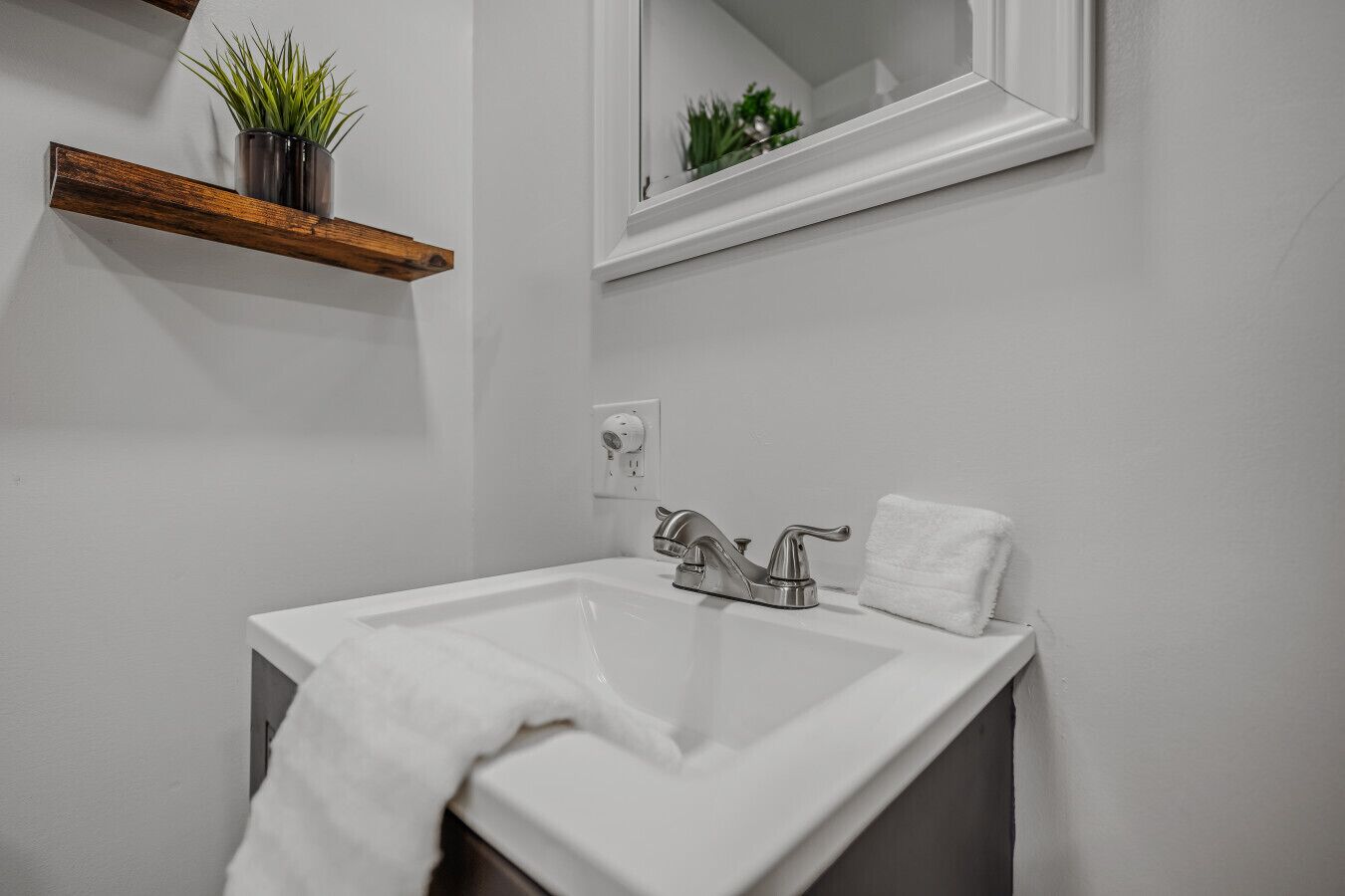 Une salle de bain avec un lavabo blanc, un miroir et une étagère en bois sur laquelle est posée une plante.
