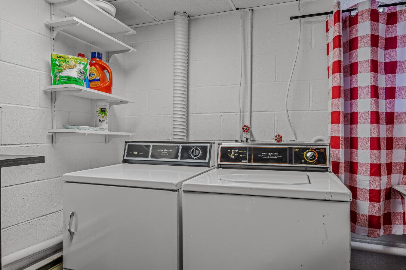 A laundry room with two white washers and dryers, a shelf with detergent, and a red and white checkered shower curtain.