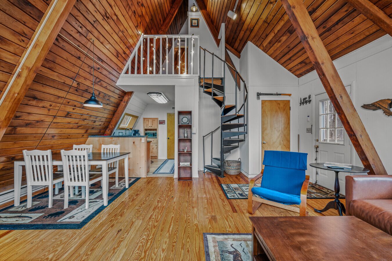 A loft-style room with a wooden ceiling, a dining area with white chairs, a blue sofa, and a spiral staircase.