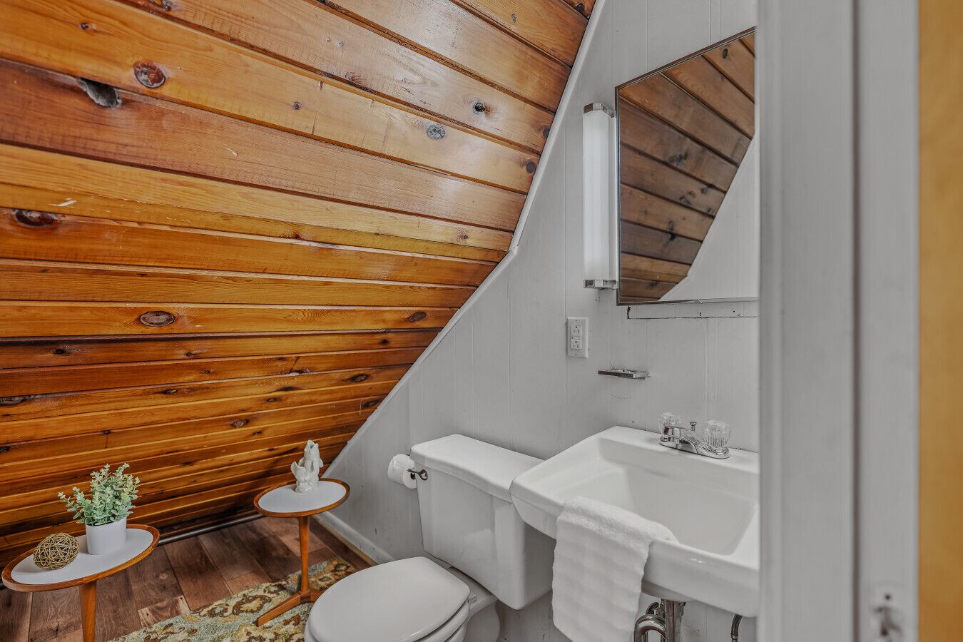 Ein kleines, rustikales Badezimmer mit Holzbalkendecke, einer weißen Toilette, einem Waschbecken mit Spiegel und einem kleinen runden Tisch mit einer Pflanze.