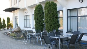An outdoor seating area with tables and chairs, surrounded by a garden with rocks and plants, and a building with large windows and a white facade. - Hotel Junior 2 (Kraków)