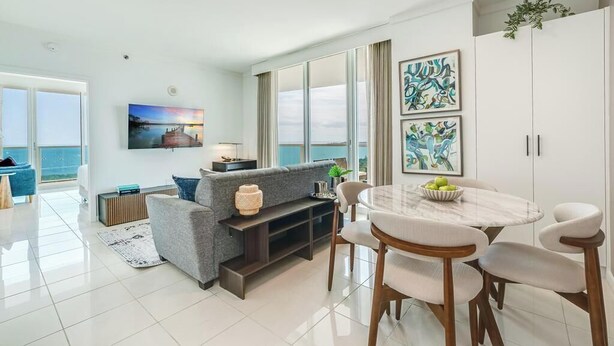 A modern living room with a grey sofa, a dining table with chairs, a flat-screen TV, and artwork on the walls.