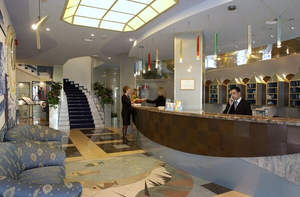 A hotel lobby with a reception desk, a staircase, and a seating area.