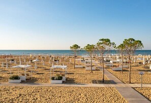 Een strand met witte ligbedden en tafels, een houten looppad en bomen.