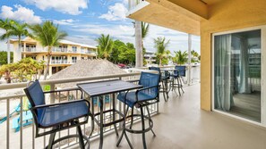 A balcony with a table and chairs, overlooking a pool area with palm trees and a building in the background.