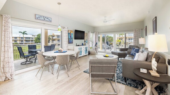 A spacious living room with a dining area, large windows, and a balcony with a view of other buildings.