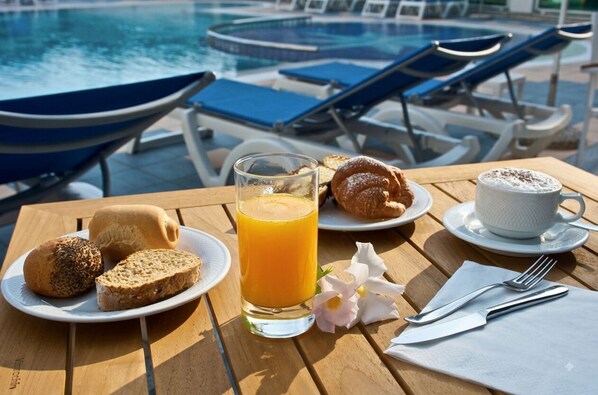A breakfast setup with croissants, bread, juice, and coffee on a wooden table by a poolside.
