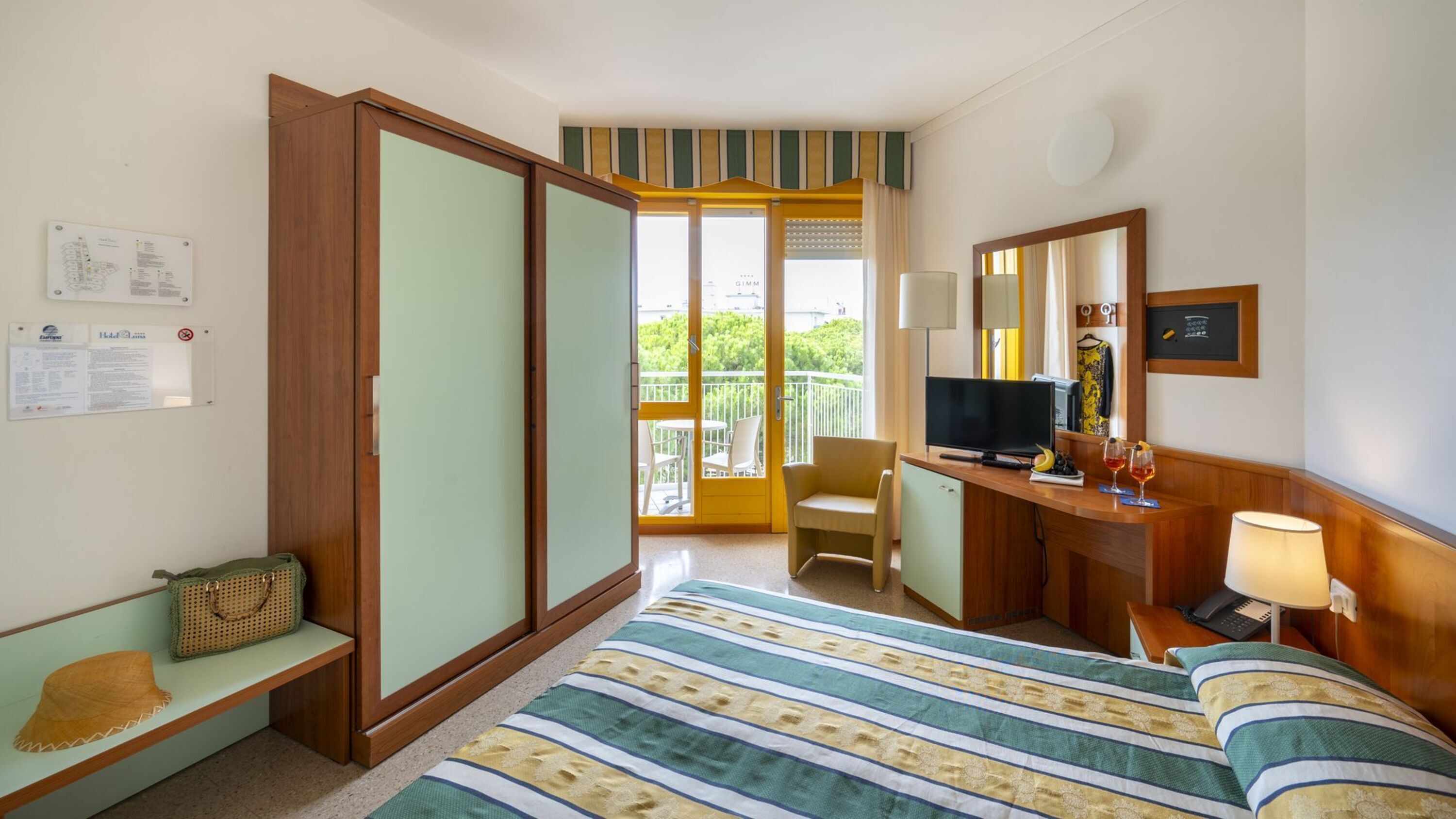 A hotel room with a bed, a desk with a television, a chair, a small table with drinks, a lamp, and a balcony with chairs.