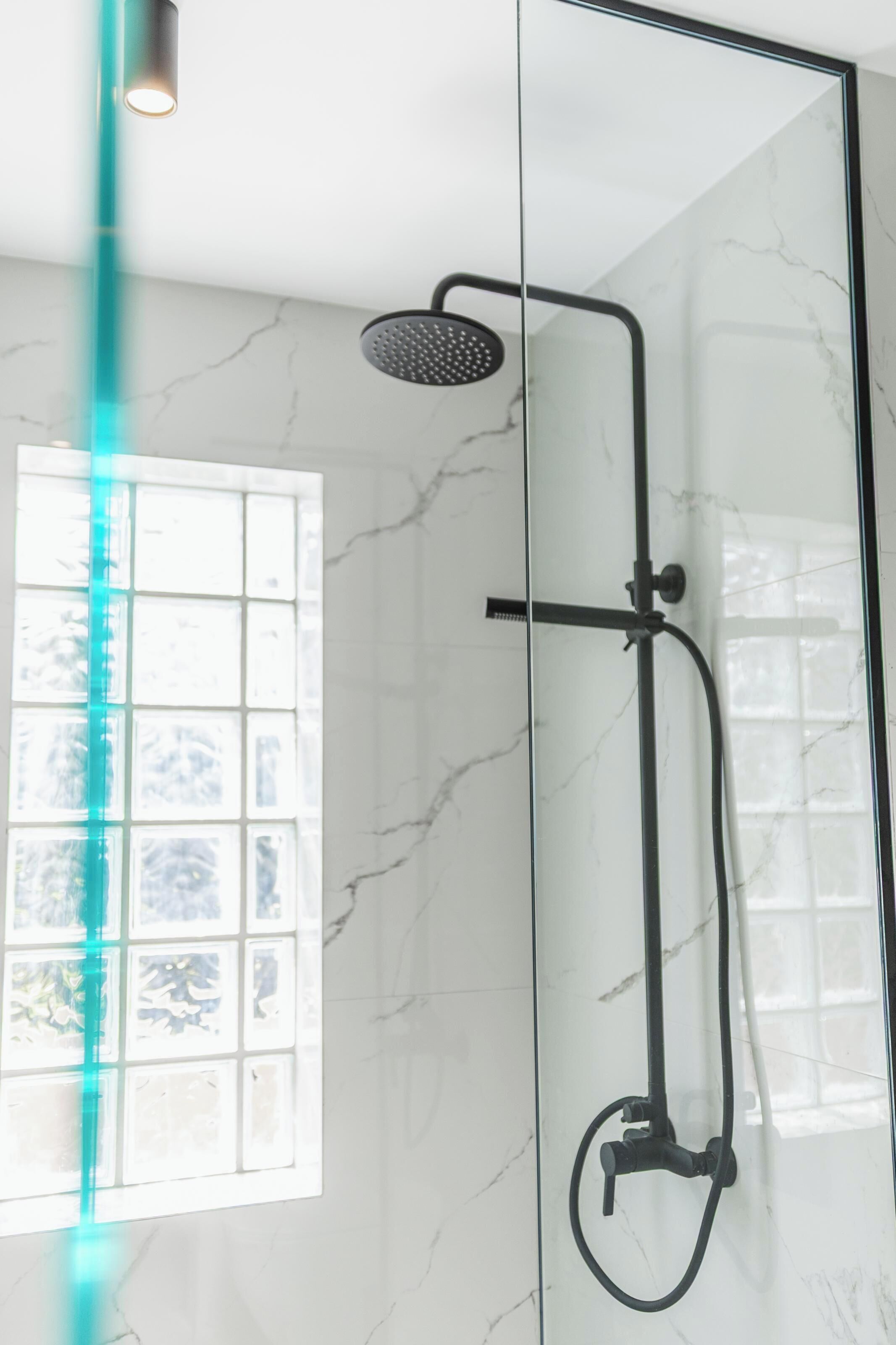 A modern bathroom with a glass shower enclosure, a visible window with a grid pattern, and a marble wall.