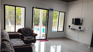 A spacious living area with a sofa set, a flat-screen TV mounted on the wall, and a view of a swimming pool through large windows.