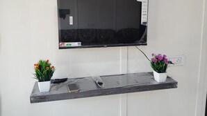A flat-screen TV mounted on a wall, with a remote control and a smartphone on a shelf below.