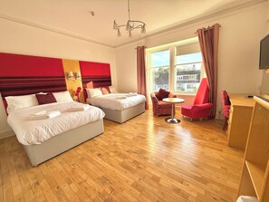 A hotel room with two beds, a small table, a red armchair, and a TV. - Seabank House Hotel (Nairn)