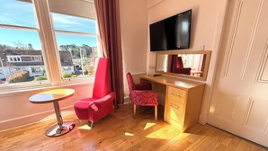 A room with a wooden floor, a red chair, a desk with a mirror, and a TV mounted on the wall. - Seabank House Hotel (Nairn)