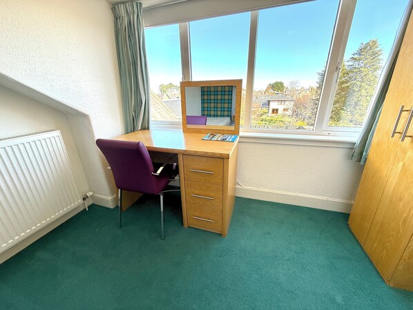 A room with a wooden desk, purple chair, and a view of buildings through large windows. - Seabank House Hotel (Nairn)