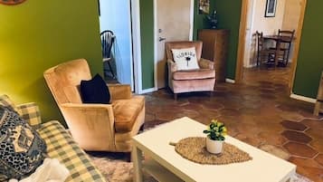 Un salon avec un canapé à carreaux, un fauteuil beige, une table basse blanche ornée d’une plante et un coussin à motifs floridiens sur une chaise.
