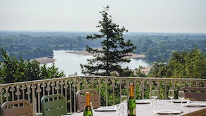 An outdoor dining area with a table set for two, overlooking a river and forested landscape. - Castle near Saumur with stunning Loire view, large nature chateau with tennis (Gennes-Val-de-Loire)
