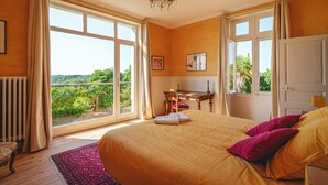 A bedroom with a large bed, a desk, and a view of greenery through the windows. - Castle near Saumur with stunning Loire view, large nature chateau with tennis (Gennes-Val-de-Loire)