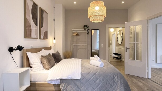 A modern bedroom with a bed, bedside lamps, a hanging light fixture, a wardrobe, and a mirror.