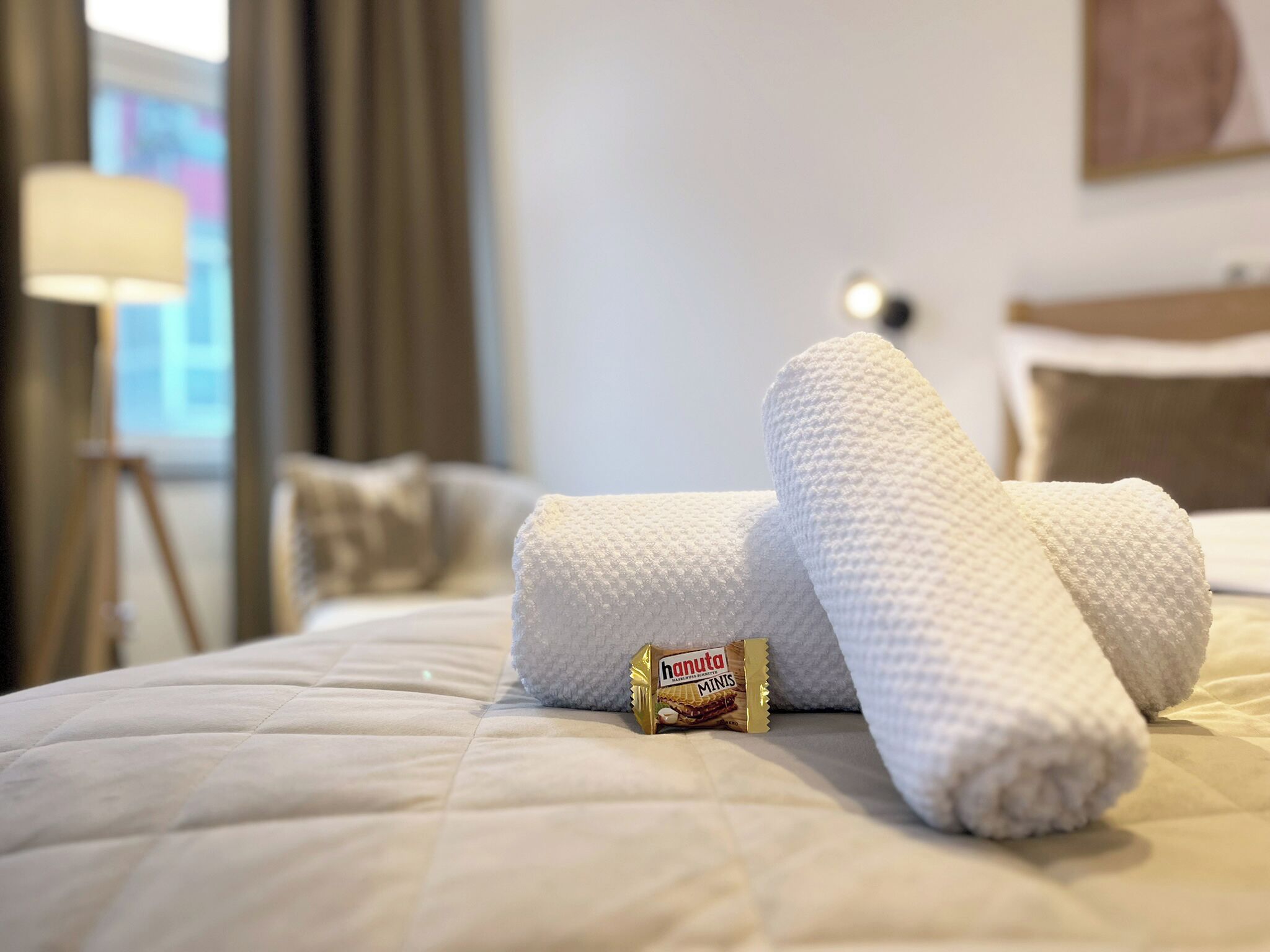 Een net opgemaakt bed met een gevouwen handdoek en een pakje Hanuta Minis chocolade.